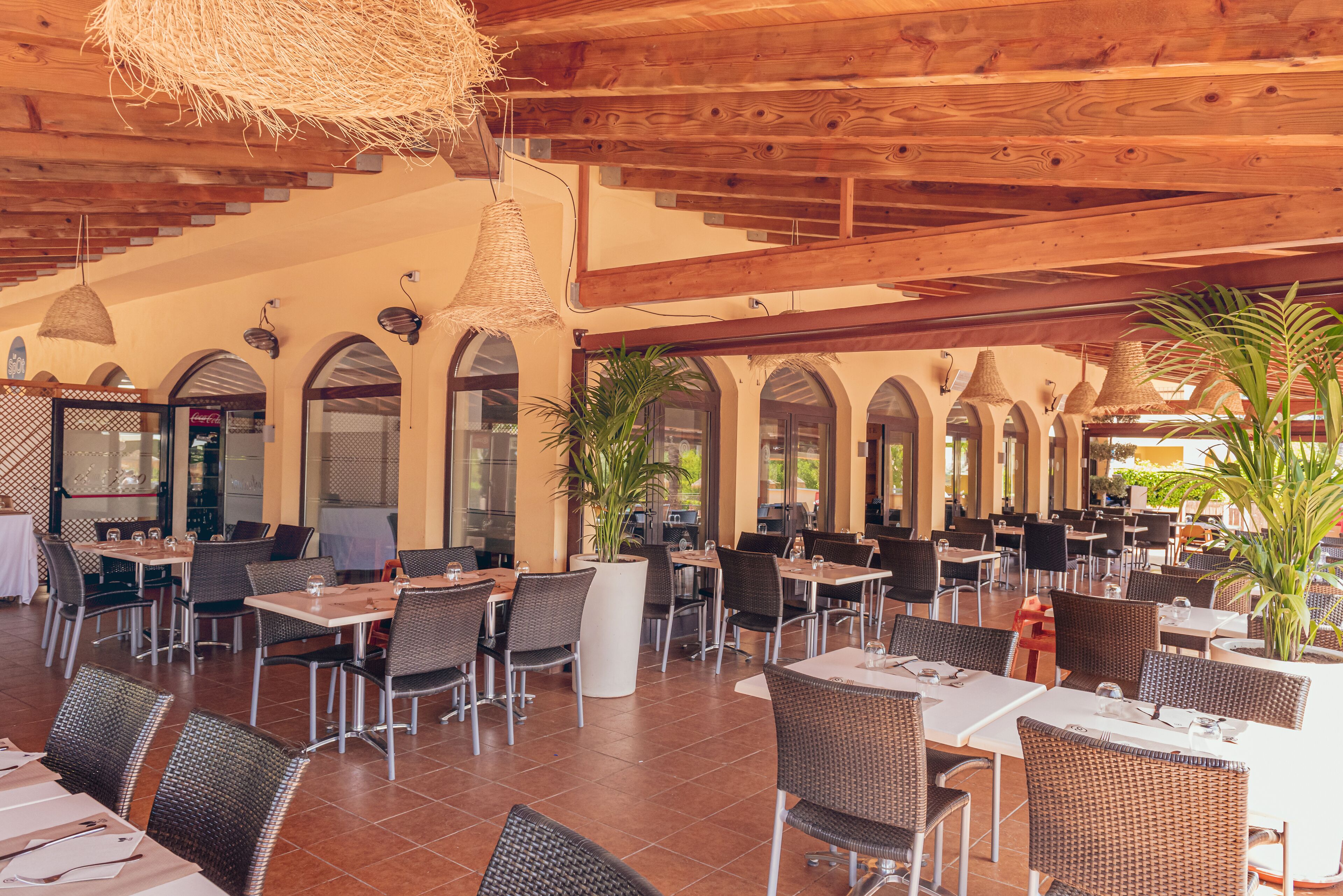An indoor dining area with multiple tables and chairs, wooden ceiling beams, and potted plants.