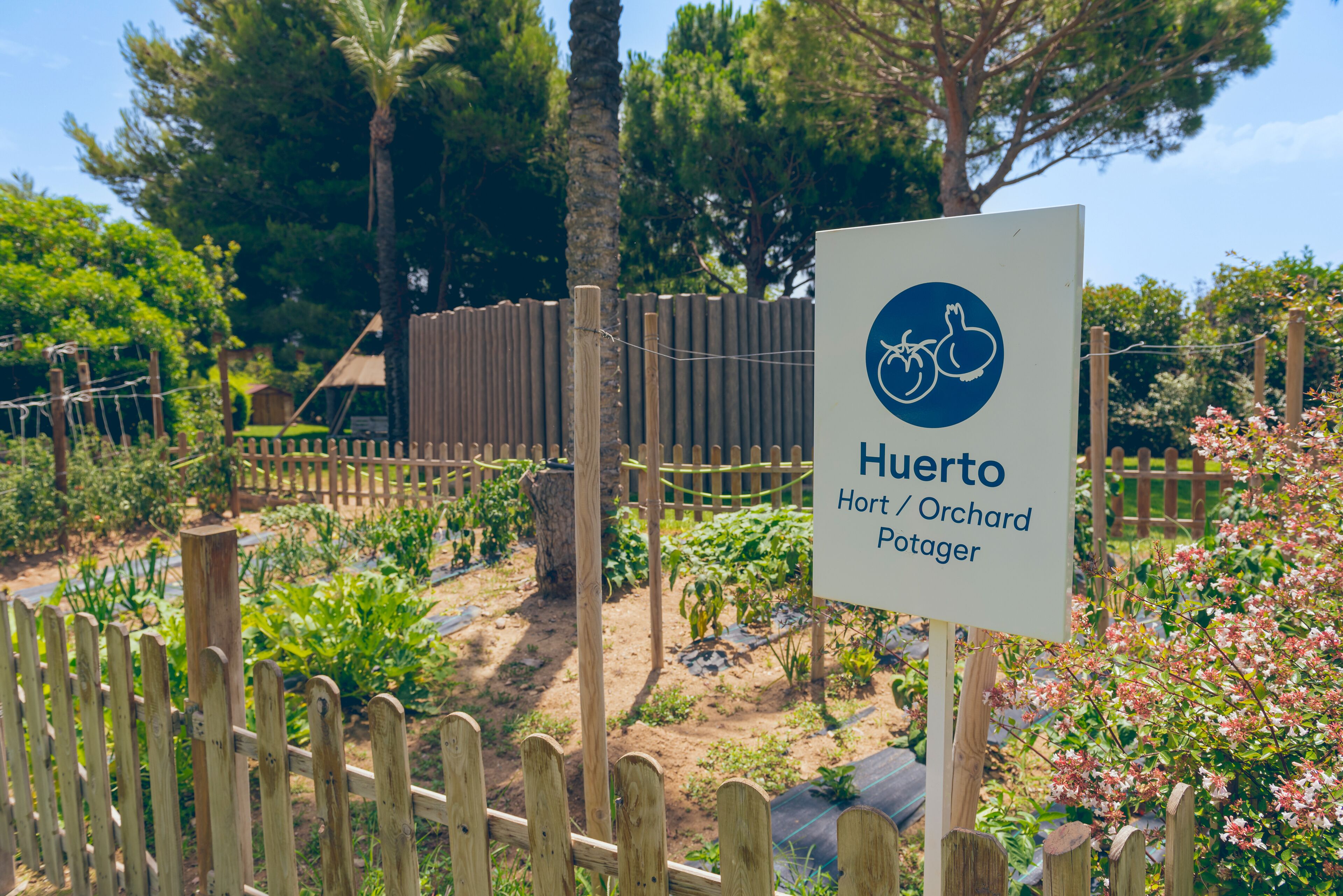 A sign for "Huerto Hort/Orchard Potager" with a wooden fence and garden in the background.