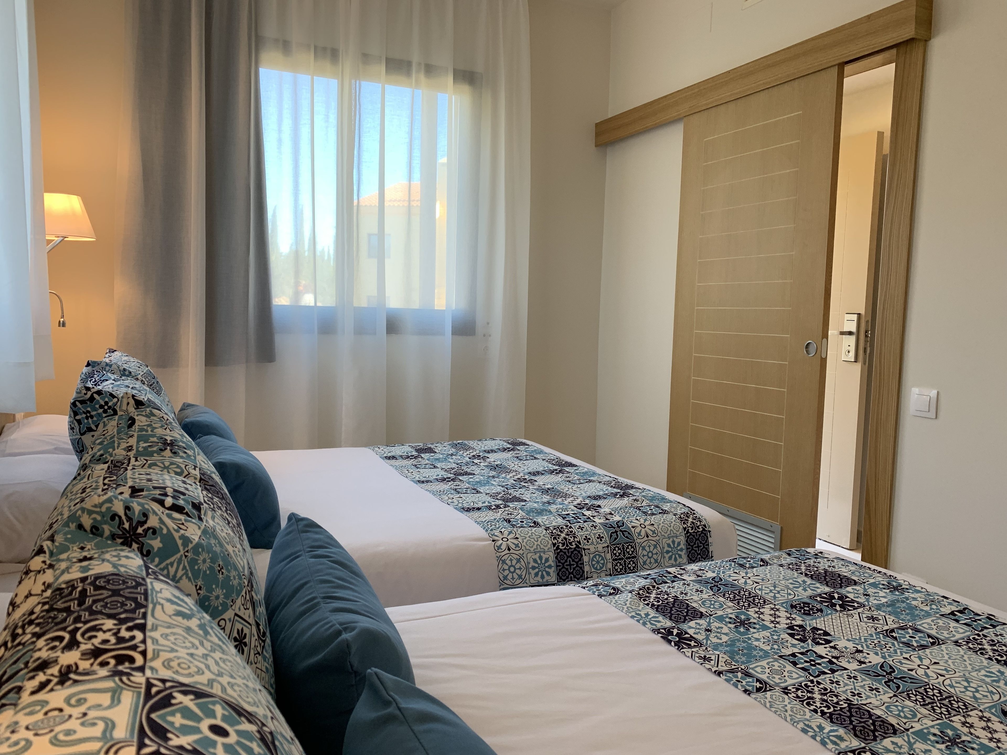 Een hotelkamer met twee bedden, een raam met dunne gordijnen, een houten deur en een lamp op een nachtkastje.