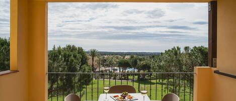 A balcony with a table set for two, overlooking a lush landscape with trees and a distant view of a resort area.