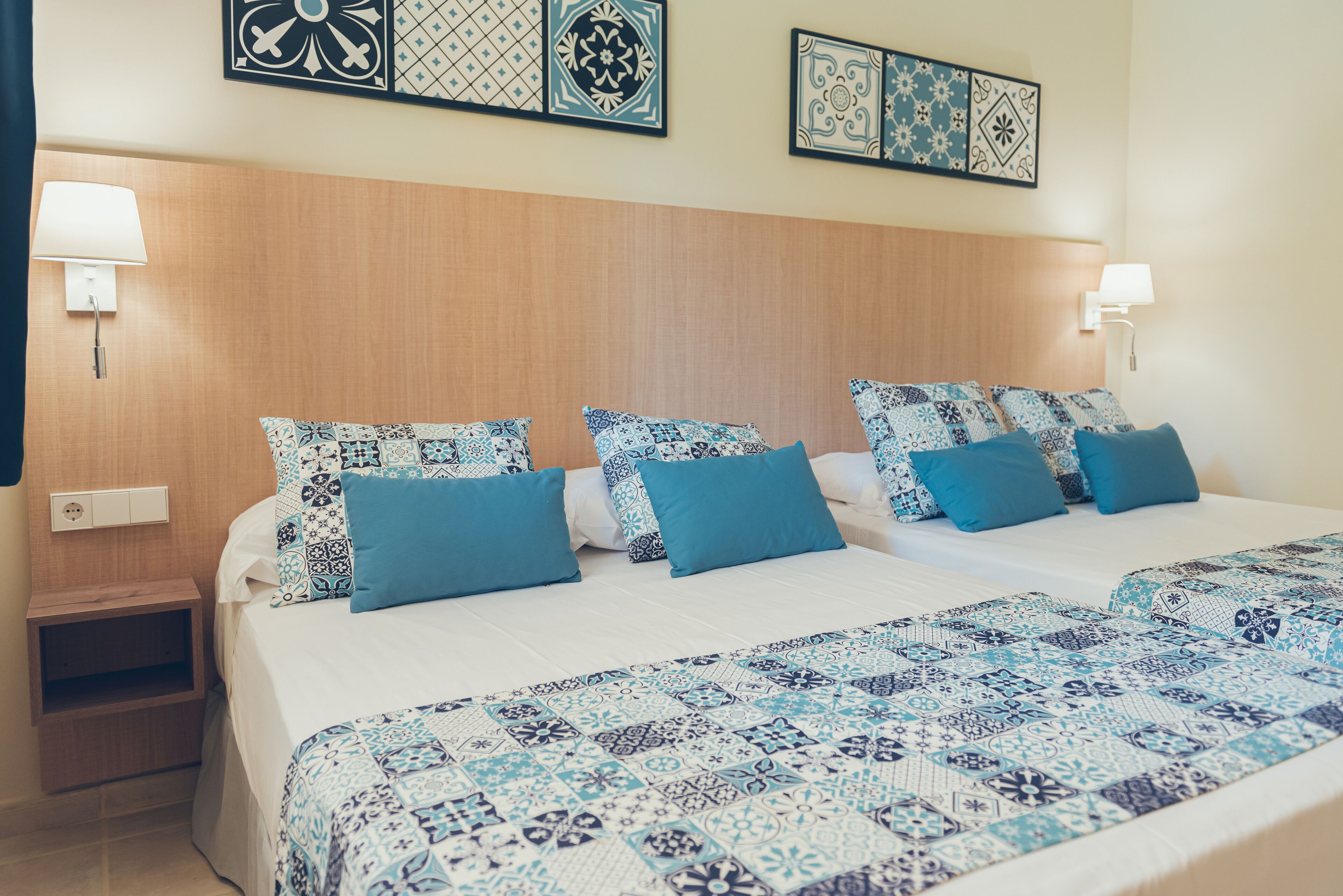 A hotel room with two beds, each with patterned bedding and matching pillows. There are wall-mounted lamps on either side of the beds, and a small wooden nightstand with a lamp.