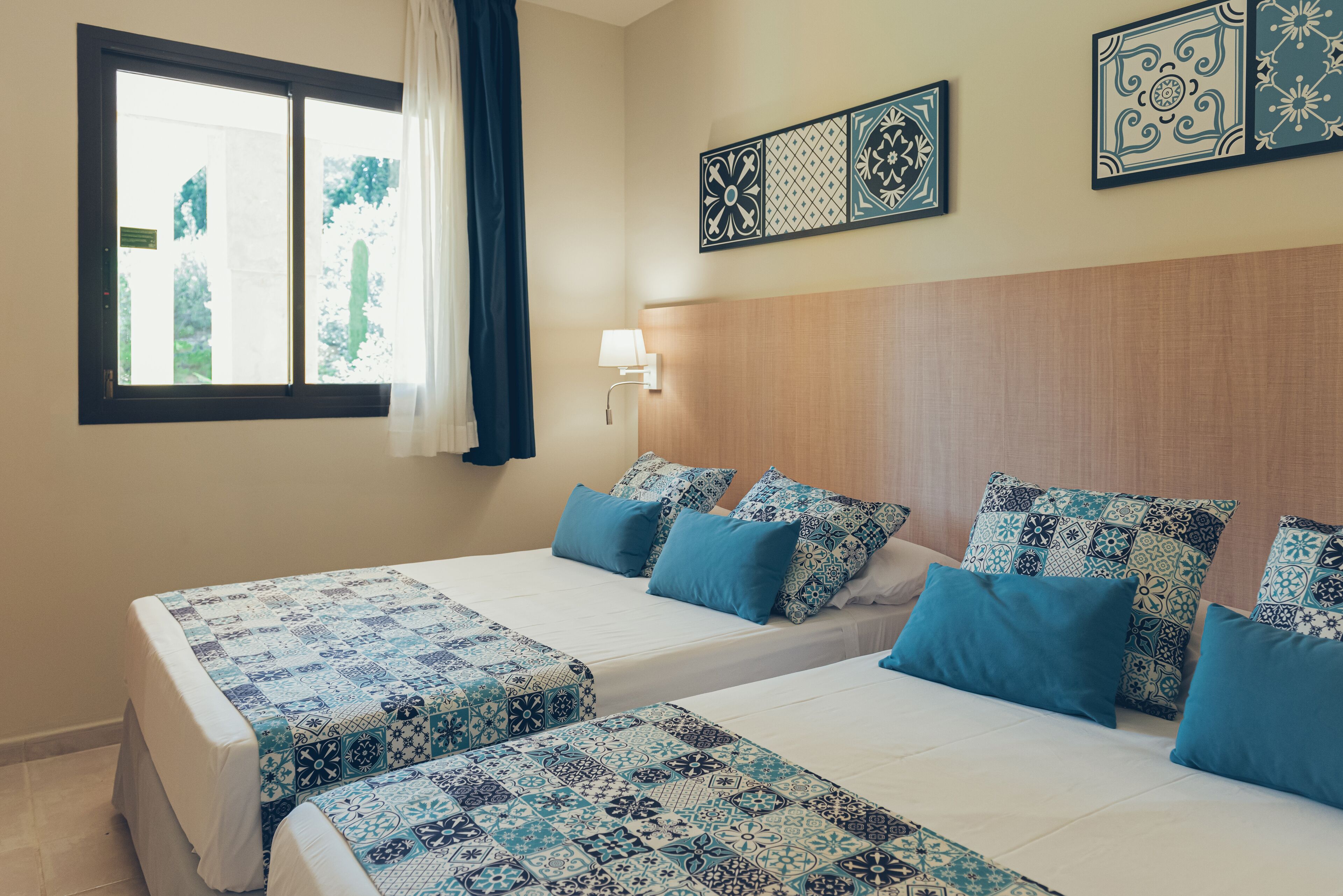 Une chambre d’hôtel avec deux lits, une fenêtre avec des rideaux, une lampe fixée au mur et des carreaux décoratifs.