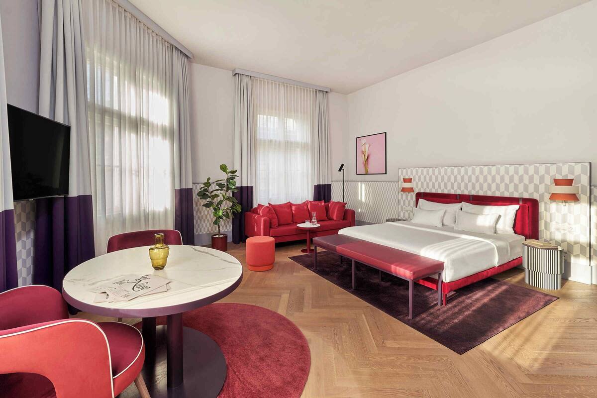 A modern hotel room with a large bed, red sofa, and a round dining table.