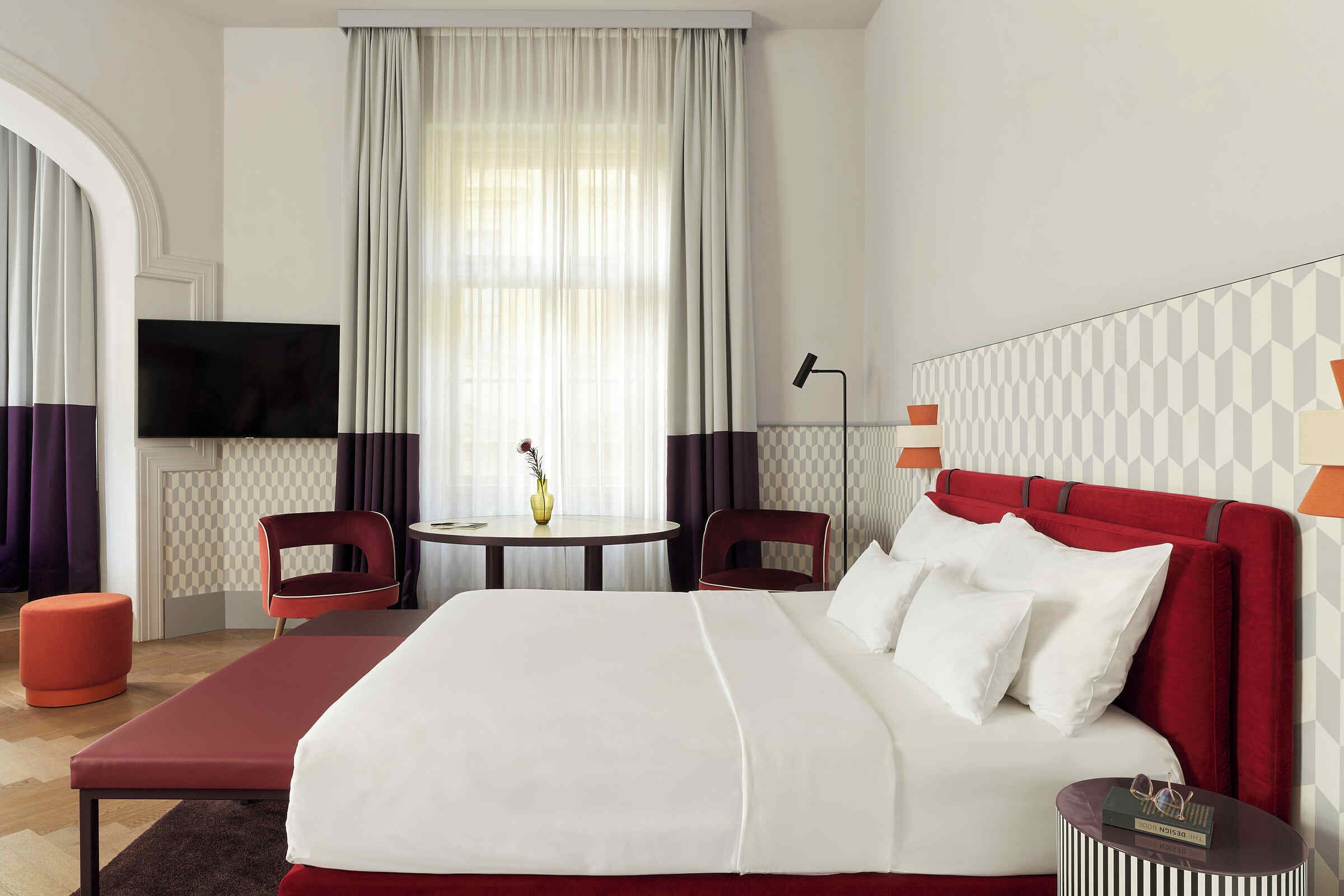 A hotel room with a large bed, a TV, a desk, two chairs, and a small table with a vase.