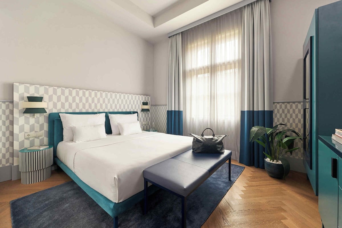 A modern hotel room with a large bed, a bedside table, a bench, a potted plant, and a wardrobe.