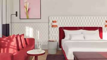A hotel room with a red sofa, a white bed with a red headboard, a small round table with a vase, and a framed picture on the wall.