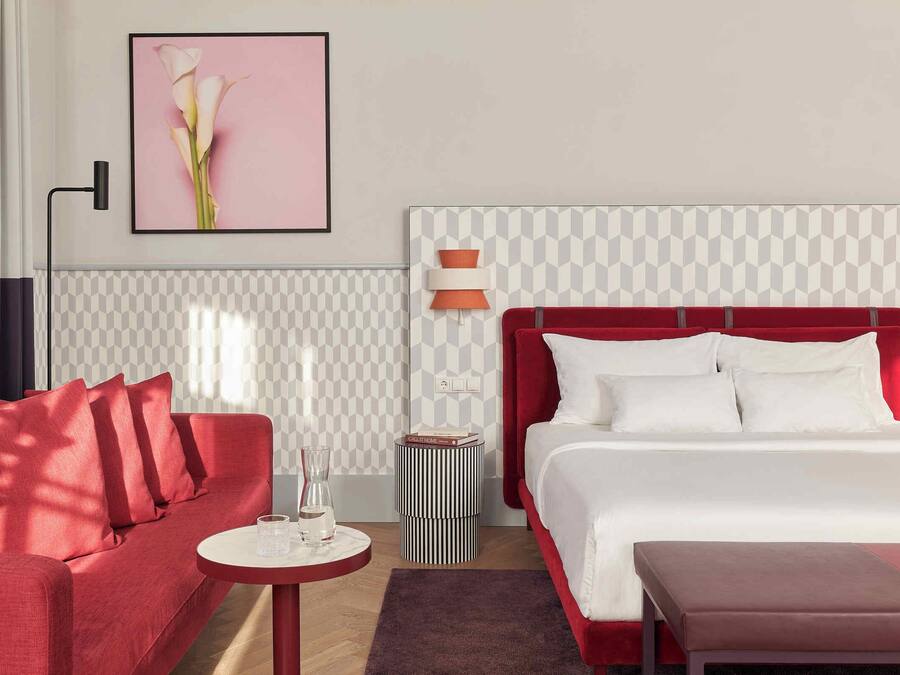 A hotel room with a red sofa, a white bed with a red headboard, a small round table with a vase, and a framed picture on the wall.
