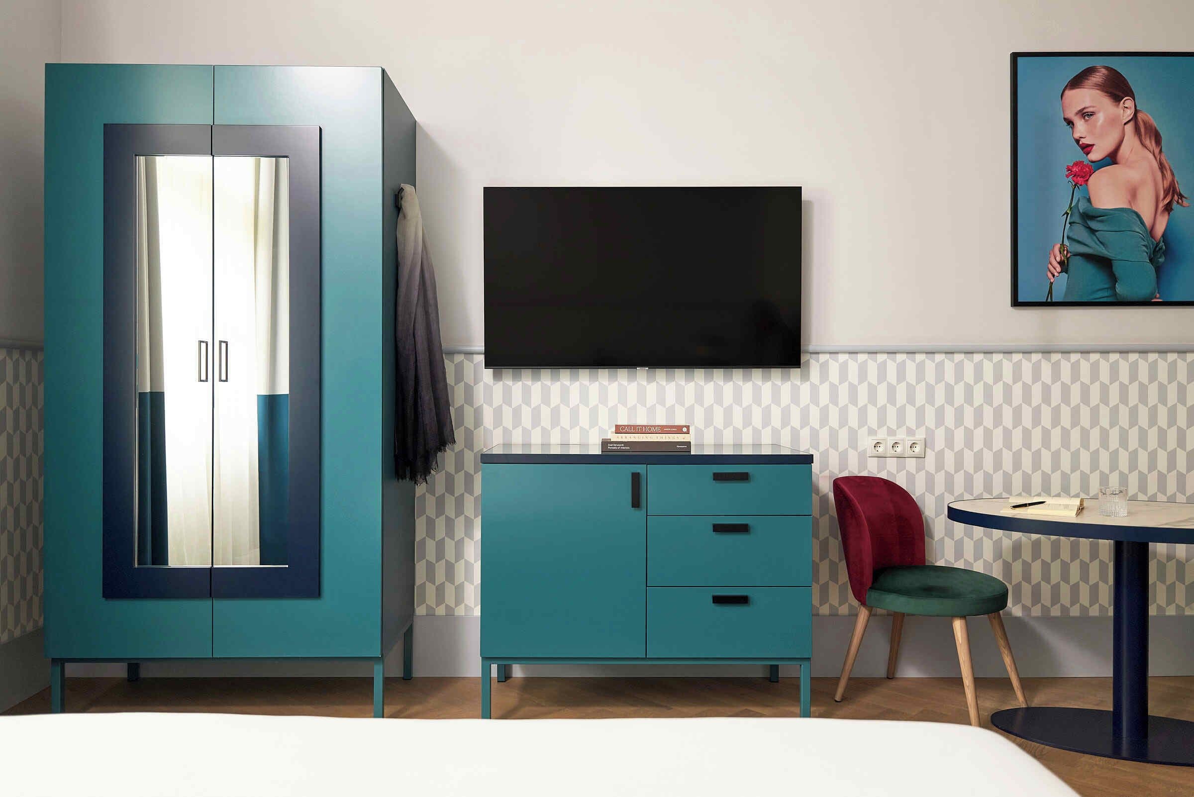 A room with a teal wardrobe, a flat-screen TV, a red chair, and a small table with a glass.