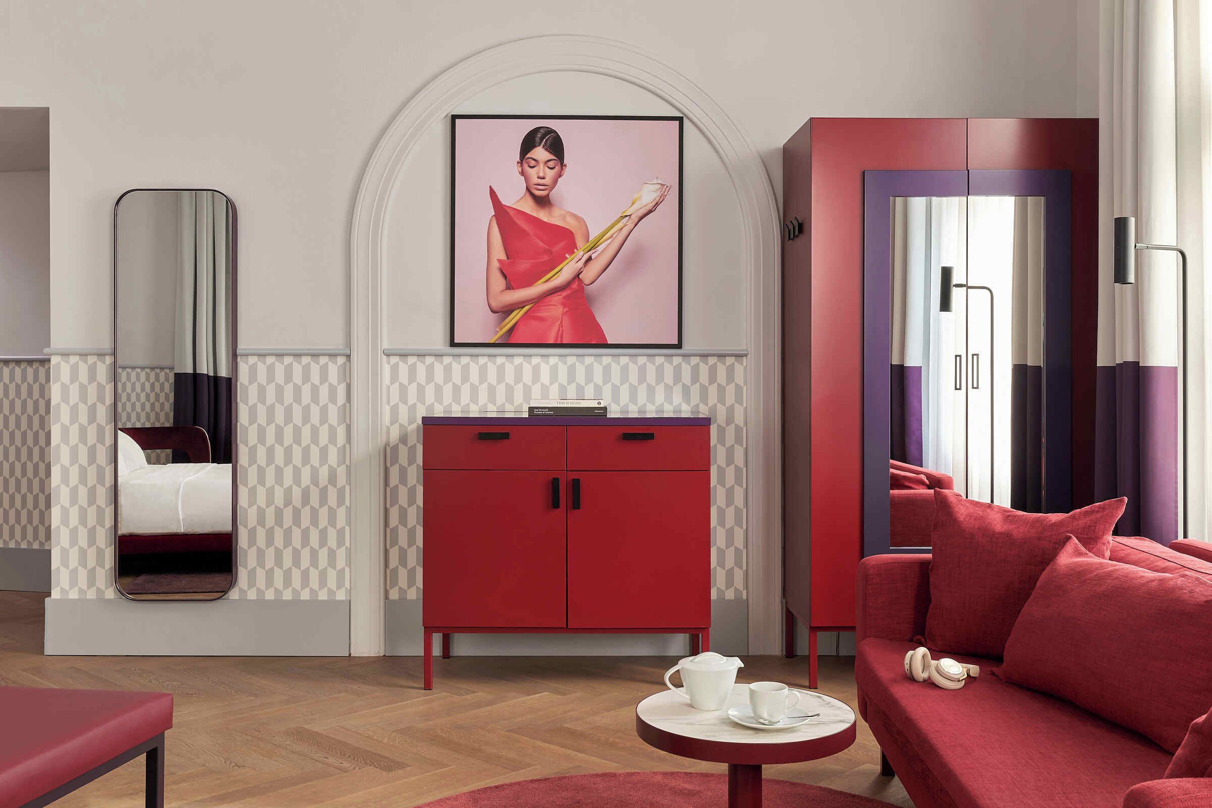 A modern hotel room with a red sofa, a small round table with a teapot, a red cabinet, and a large mirror.