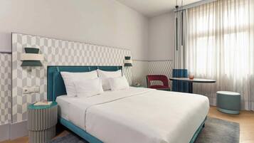 Quarto de hotel moderno com uma cama grande, uma mesa de cabeceira, uma cadeira e uma pequena mesa com uma luminária.