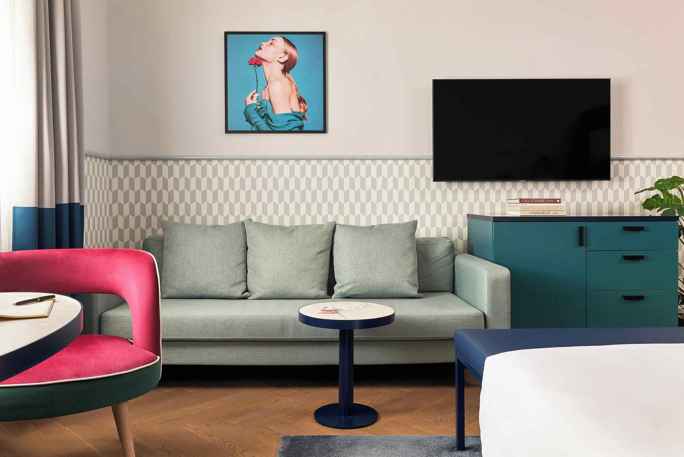 Una habitación de hotel moderna con sofá, una mesita redonda, una silla roja, una cama, un televisor y una estantería.