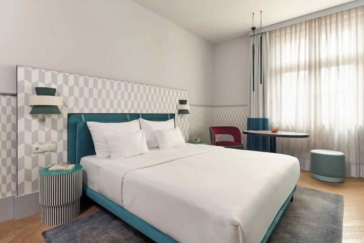 A modern hotel room with a large bed, a bedside table, a small table, and a chair.