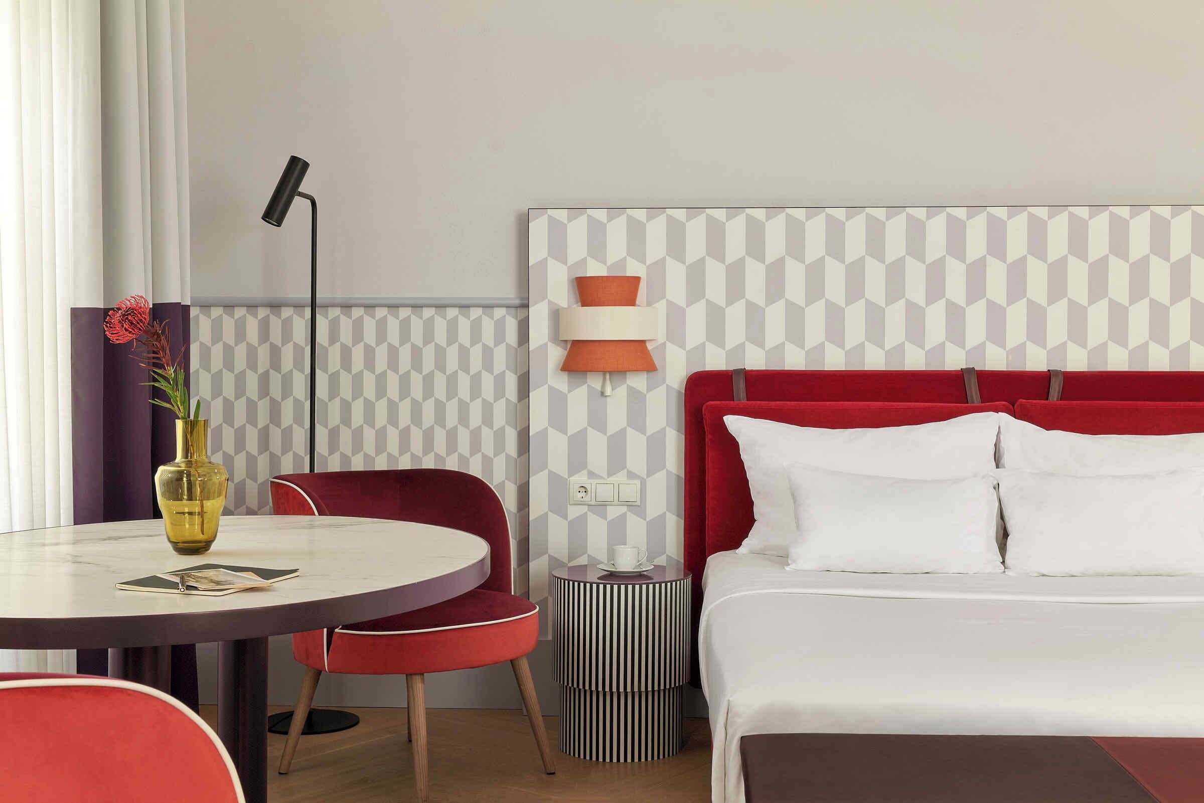 Habitación de hotel con una mesa de comedor redonda, una silla roja, una cama con ropa de cama blanca y una lámpara fijada a la pared.