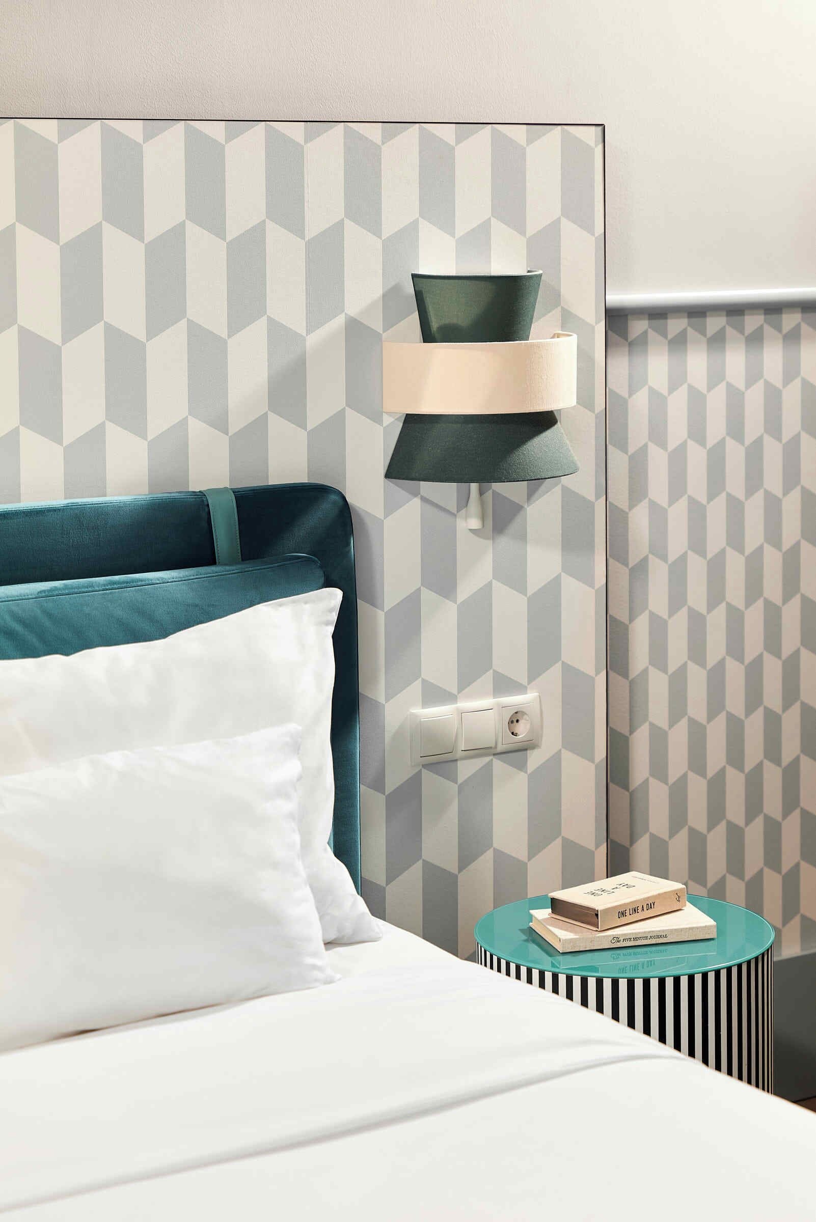 A neatly made bed with white bedding and teal pillows, a modern wall with a geometric pattern, a small round side table with a lamp, and a book titled "One Line a Day".