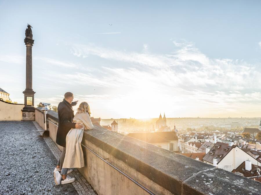 A couple on a stone ledge overlooking a cityscape with a prominent monument and a church in the distance.