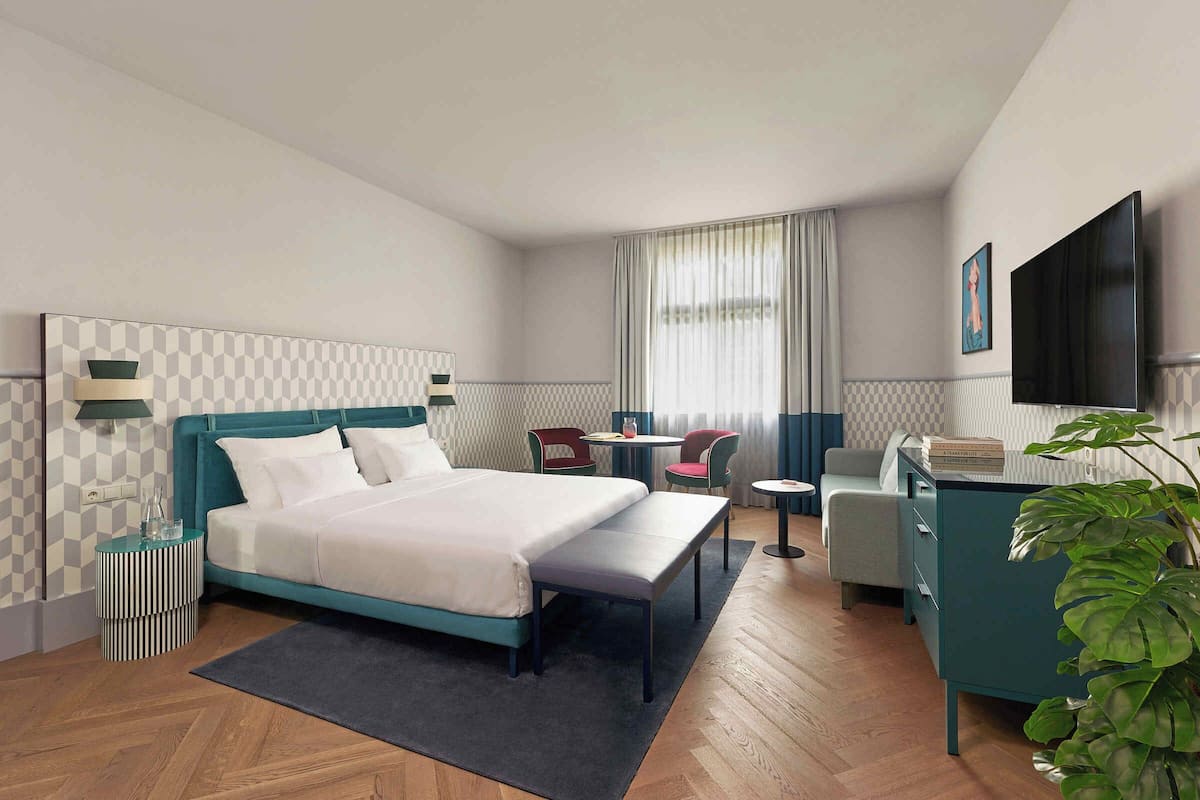 A modern hotel room with a large bed, a desk, a chair, a TV, and a plant.