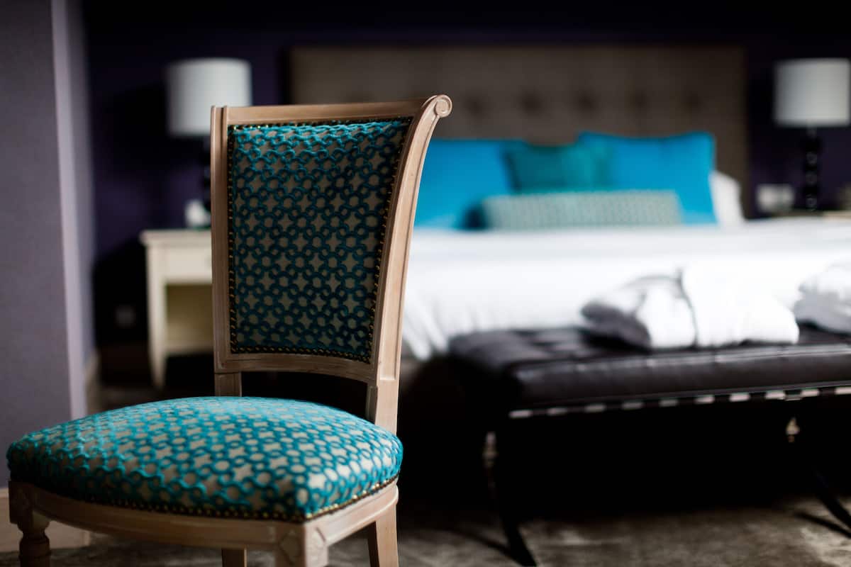 A hotel room with a wooden chair with a patterned cushion, a bed with blue and white bedding, and a black leather bench.