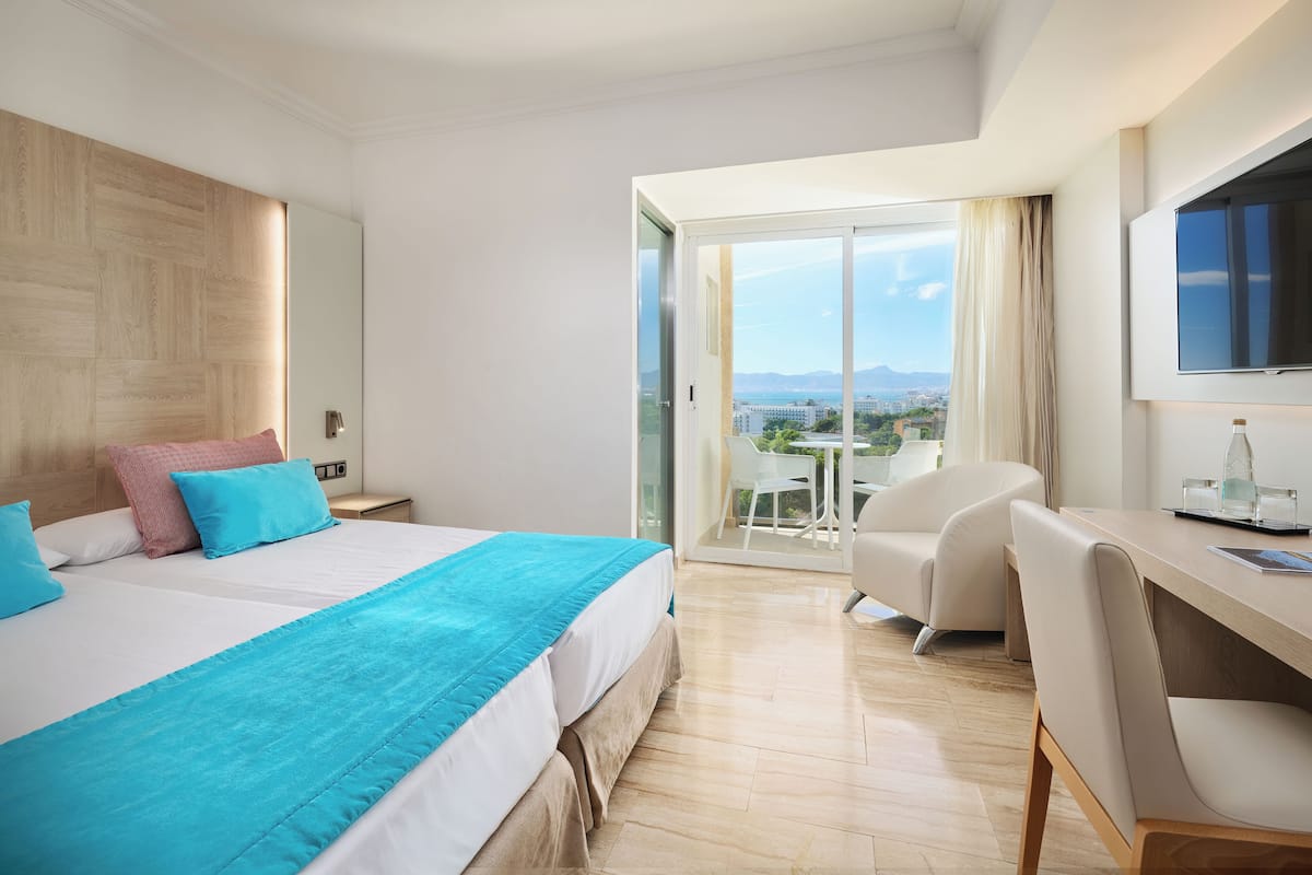 A modern hotel room with a large bed, a desk, and a balcony with a city view.