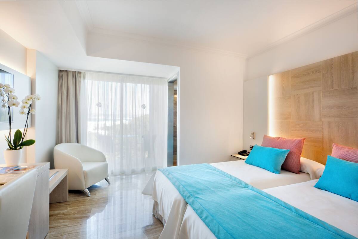 A hotel room with a bed, bedside table, chair, and a desk with a plant.