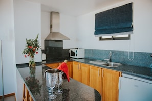 A kitchen with wooden cabinets, a black countertop, a microwave, and a sink. - Casa da Capelinha (São Vicente)