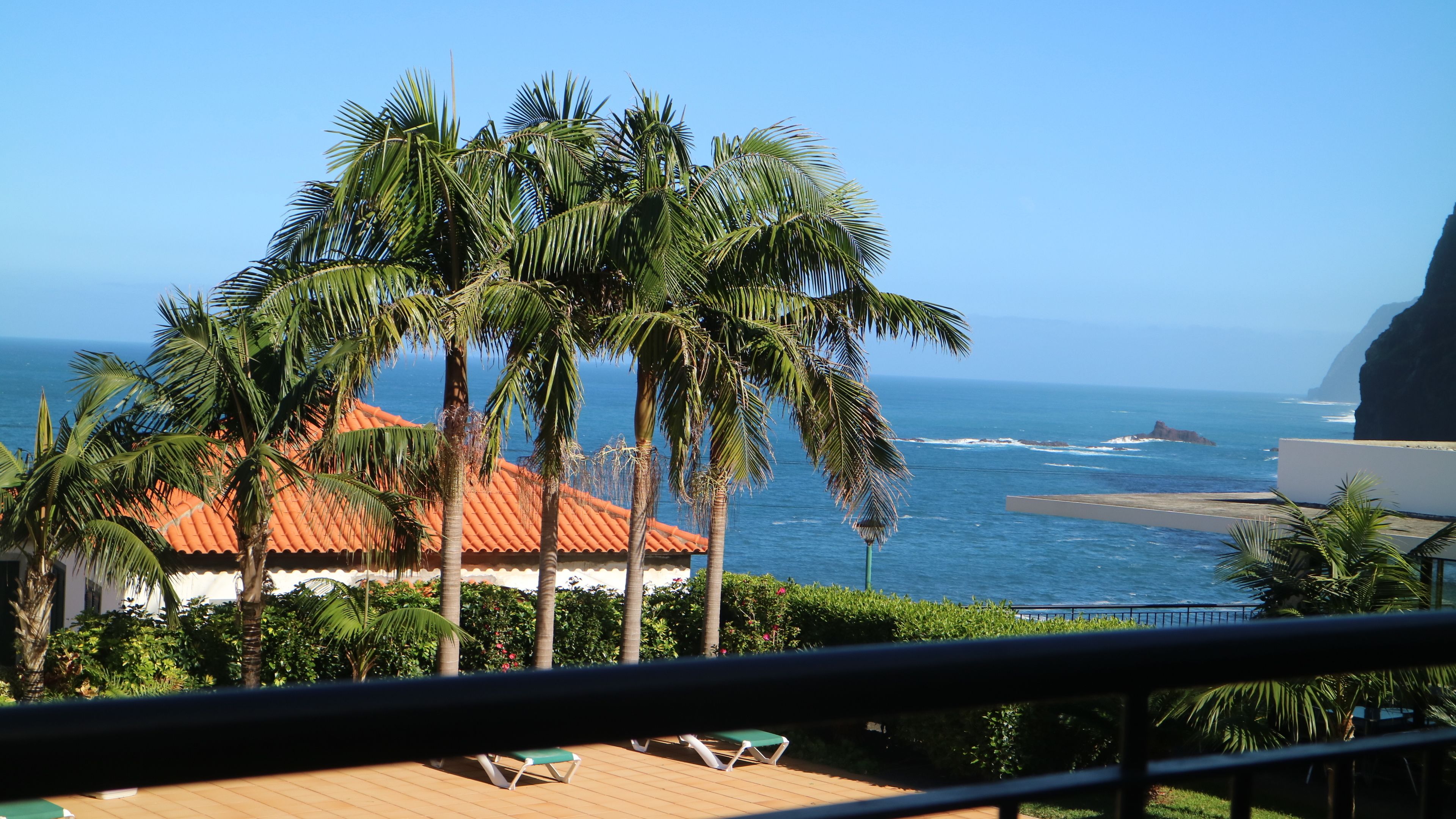 Een balkon met uitzicht op de oceaan, palmbomen en een gebouw met een rood dak.