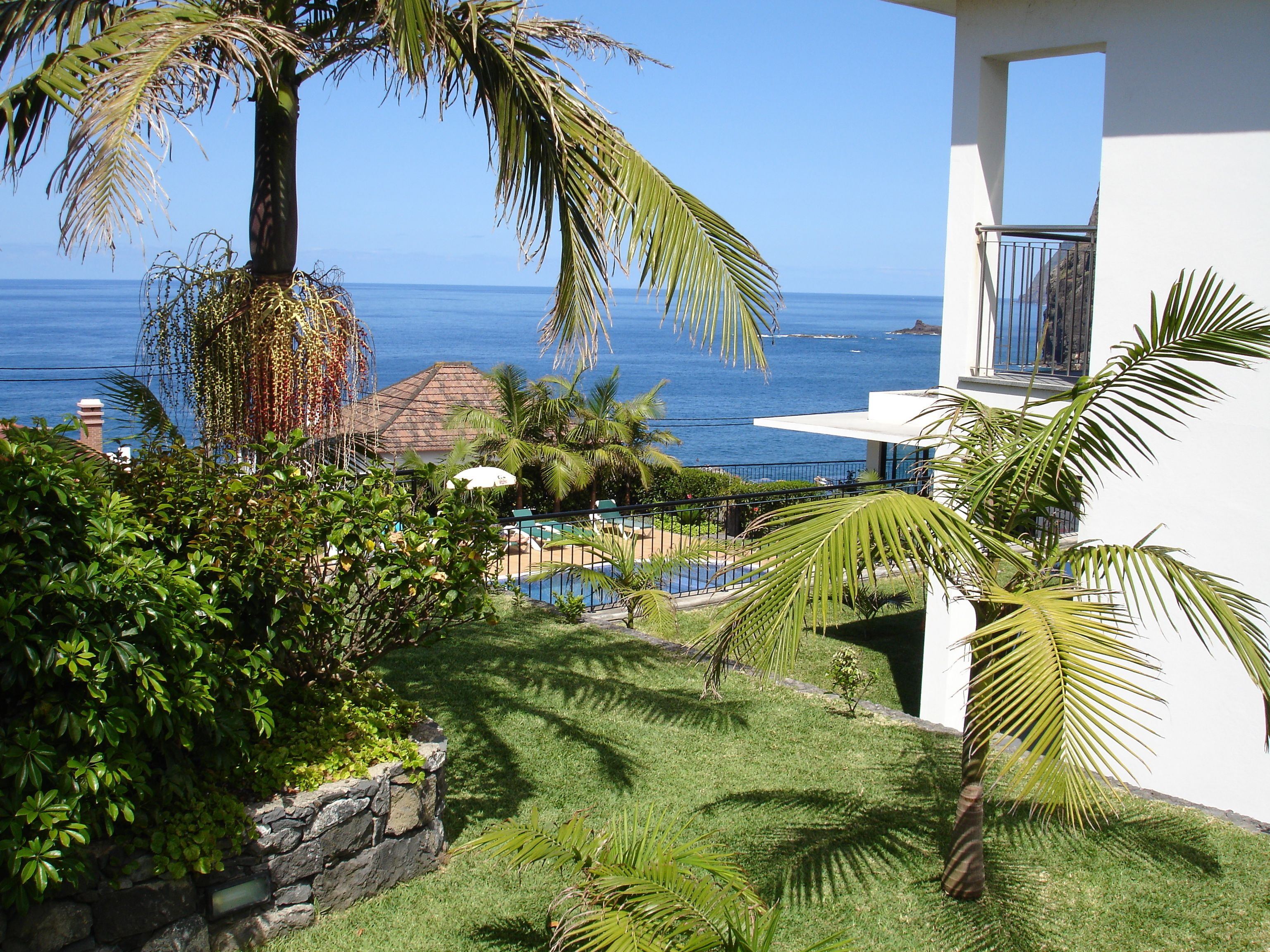A pool area with a view of the ocean, surrounded by lush greenery and a white building with a balcony.