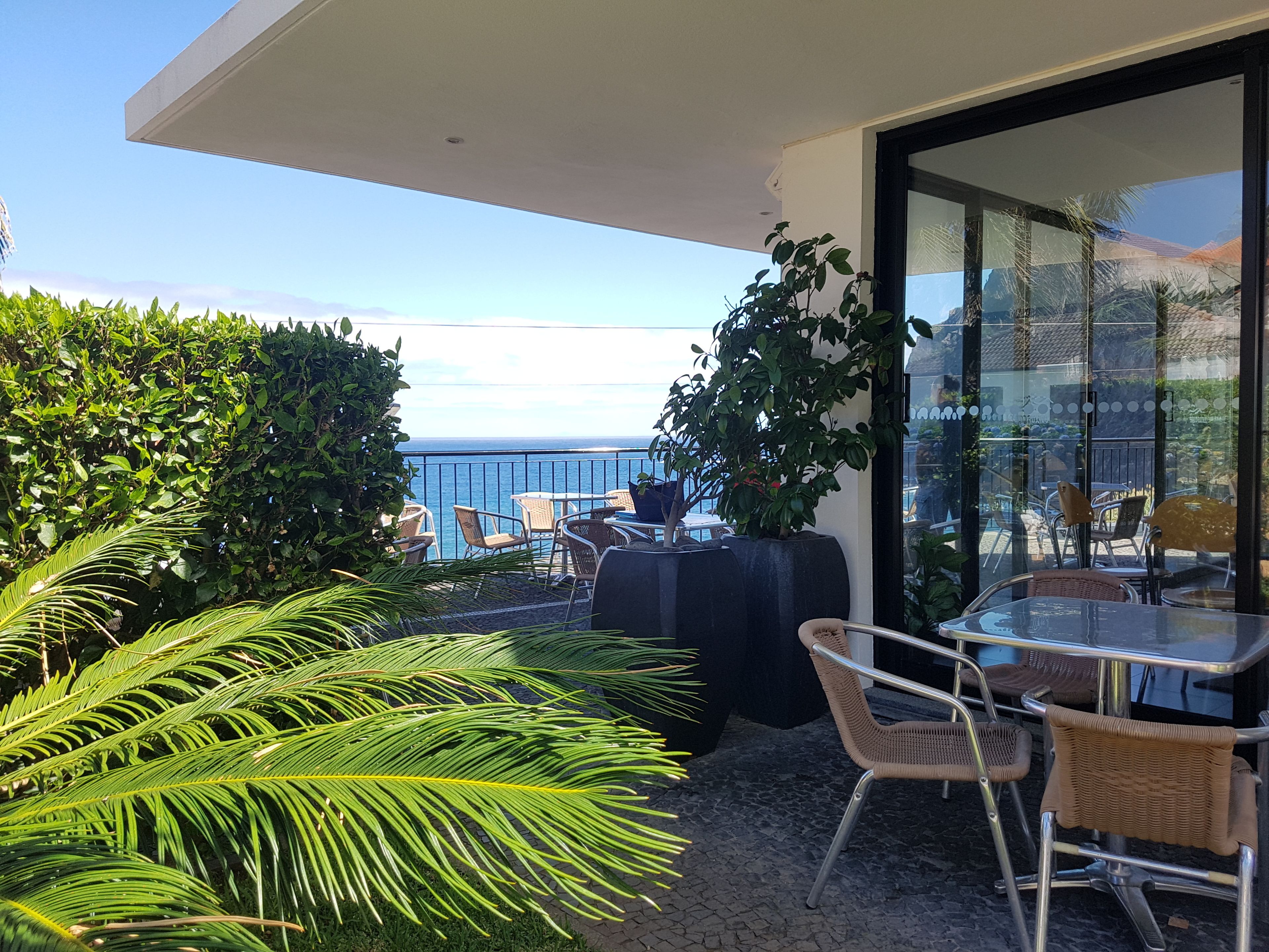 A balcony with a view of the ocean, furnished with wicker chairs and a table, surrounded by greenery.