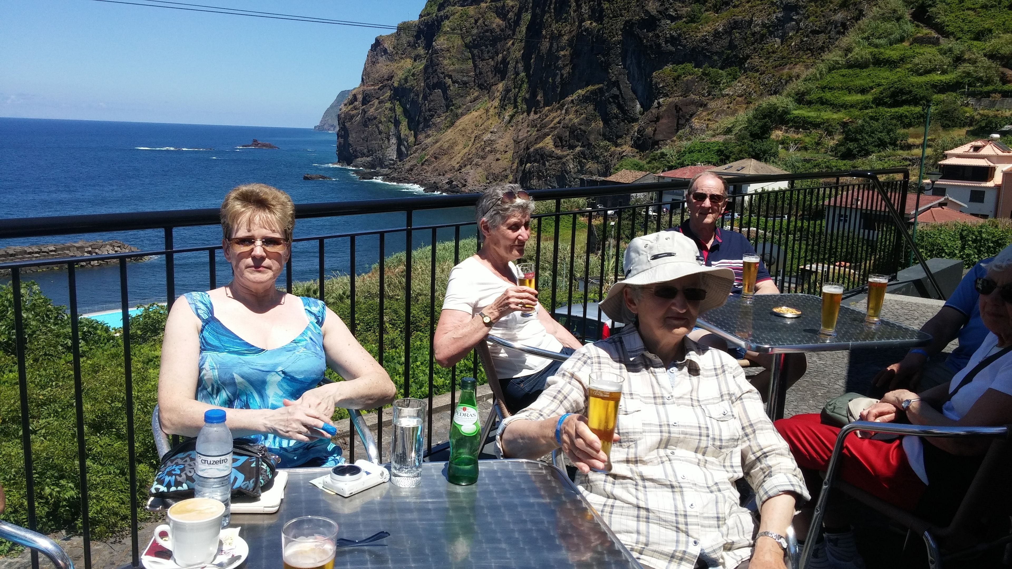 Un groupe de personnes est assis à une table à l’extérieur, devant des boissons, et profite de la vue sur une falaise côtière et l’océan.