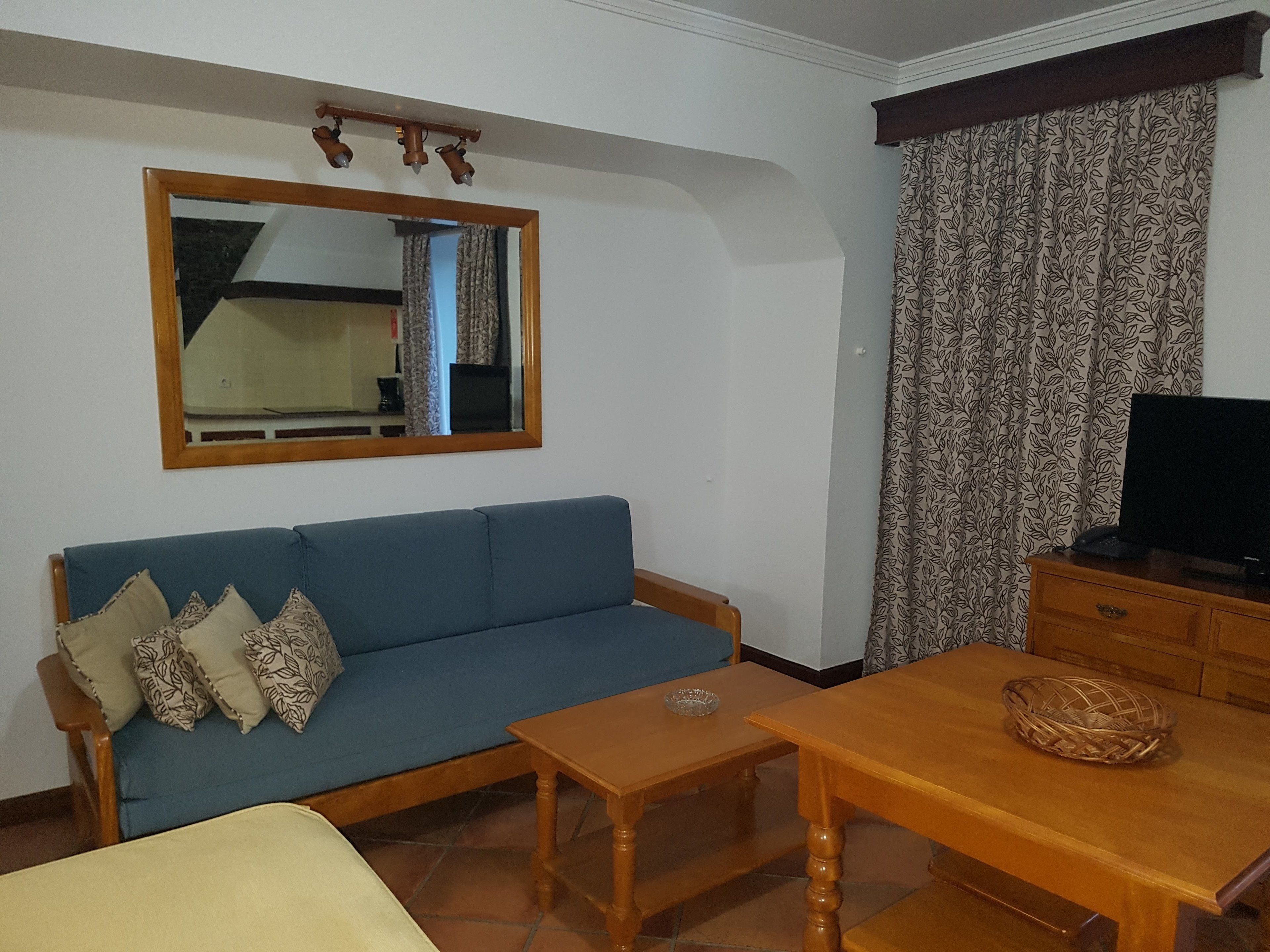 A living room with a blue sofa, a wooden coffee table, a TV on a wooden cabinet, and patterned curtains.