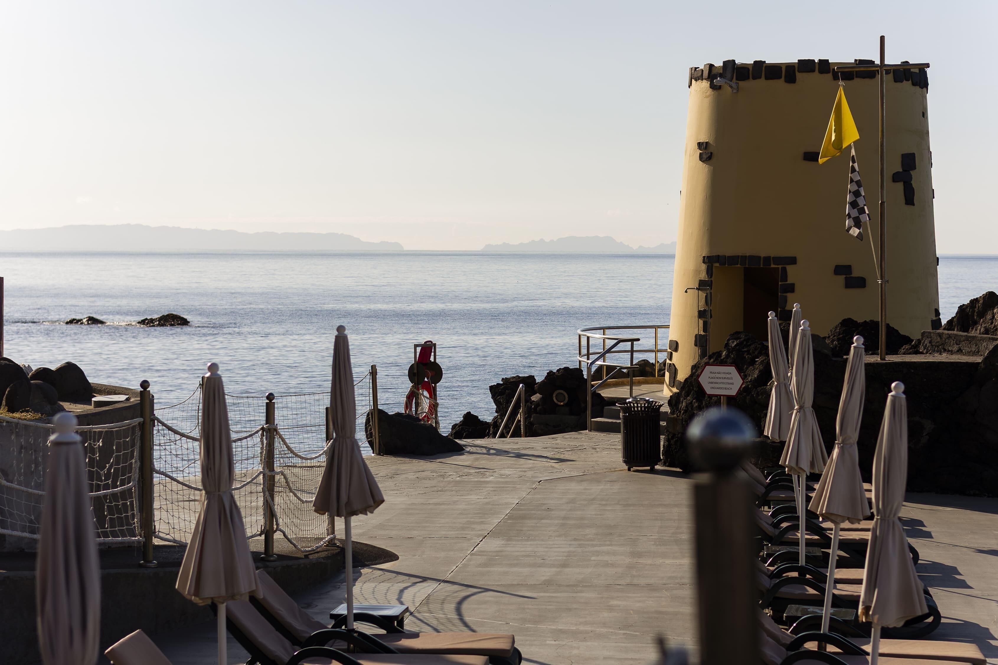 Un hotel frente al mar con una terraza de concreto, sillas de descanso, sombrillas y una estructura tipo torre.