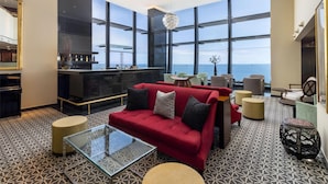 A modern lounge area with a red sofa, round tables, and a bar. Large windows offer a view of the sea. - Royal Savoy - Ocean Resort - Savoy Signature (Funchal)