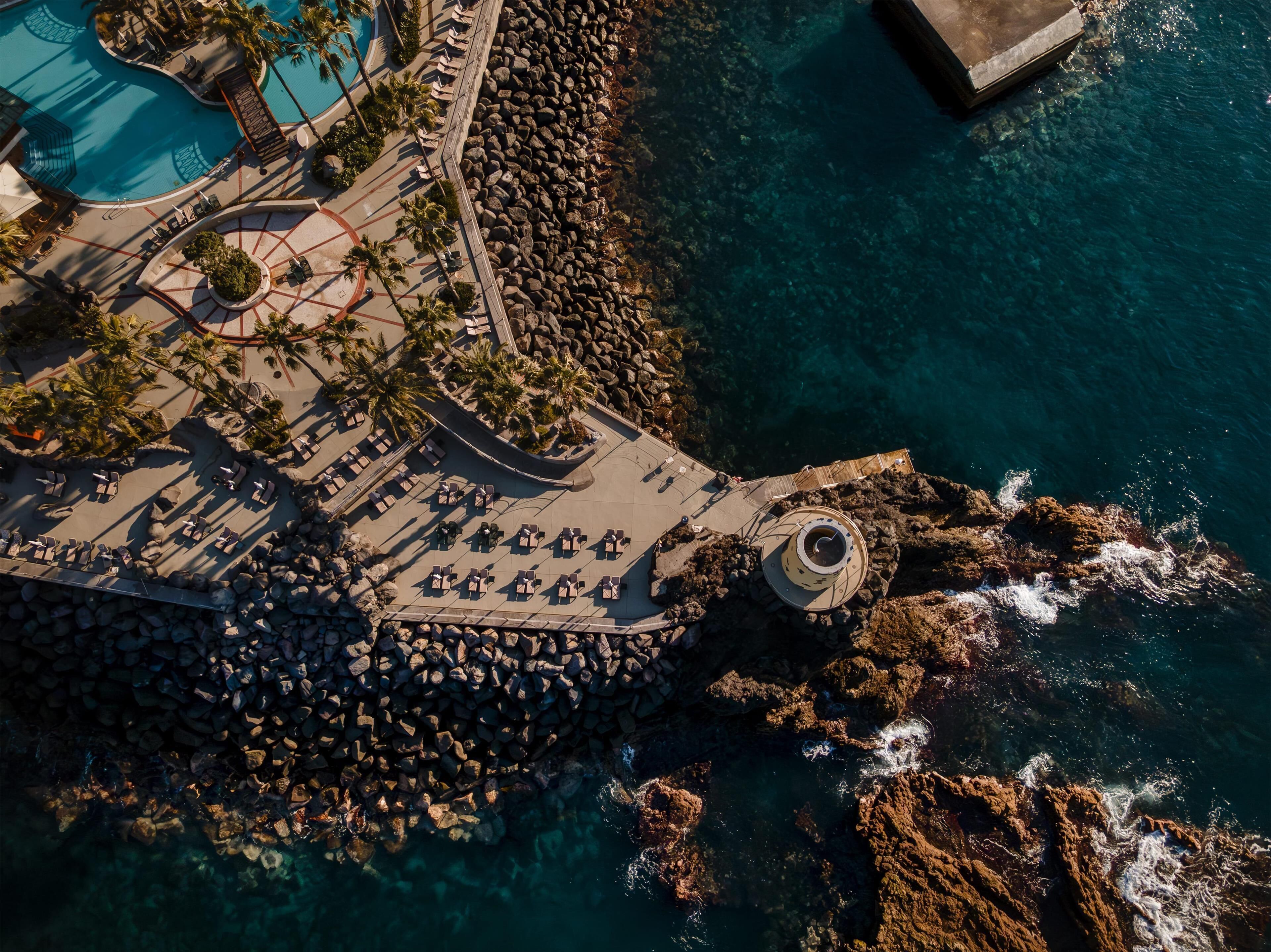 Vista aérea de um resort à beira-mar, com piscina, áreas de lazer e costa rochosa.