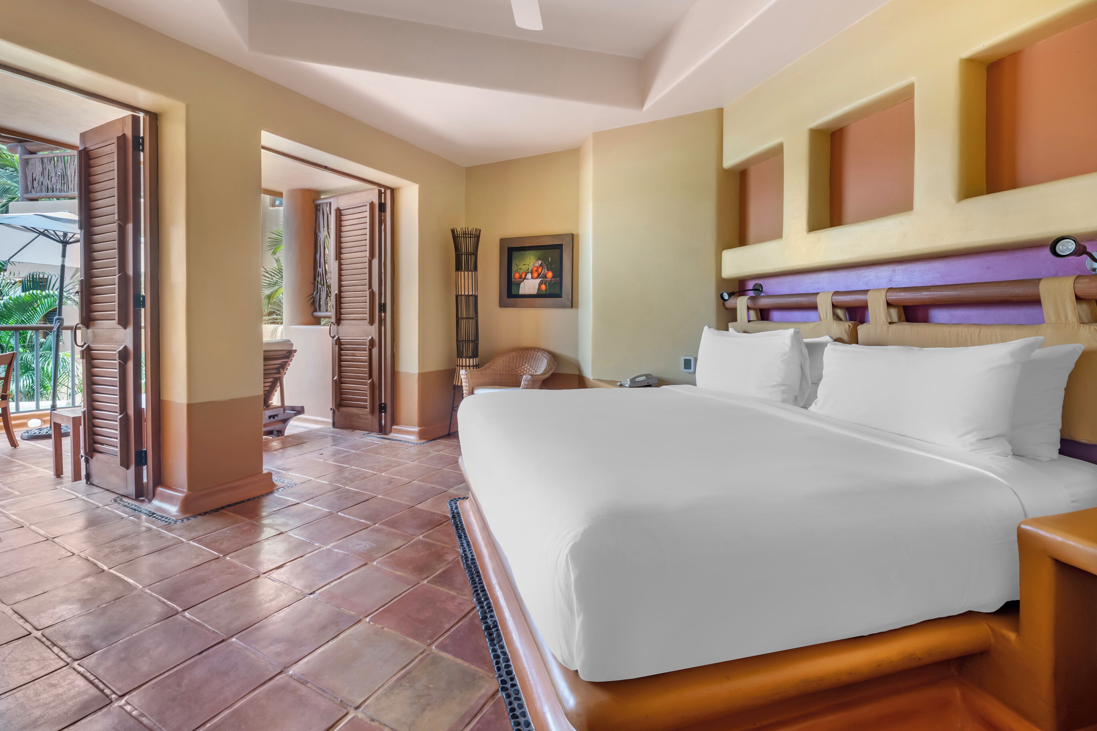 Habitación de hotel con una cama grande, cabecera de madera, una mesita y una silla. Se ve una puerta que da a un espacio al aire libre con una mesa y sillas.
