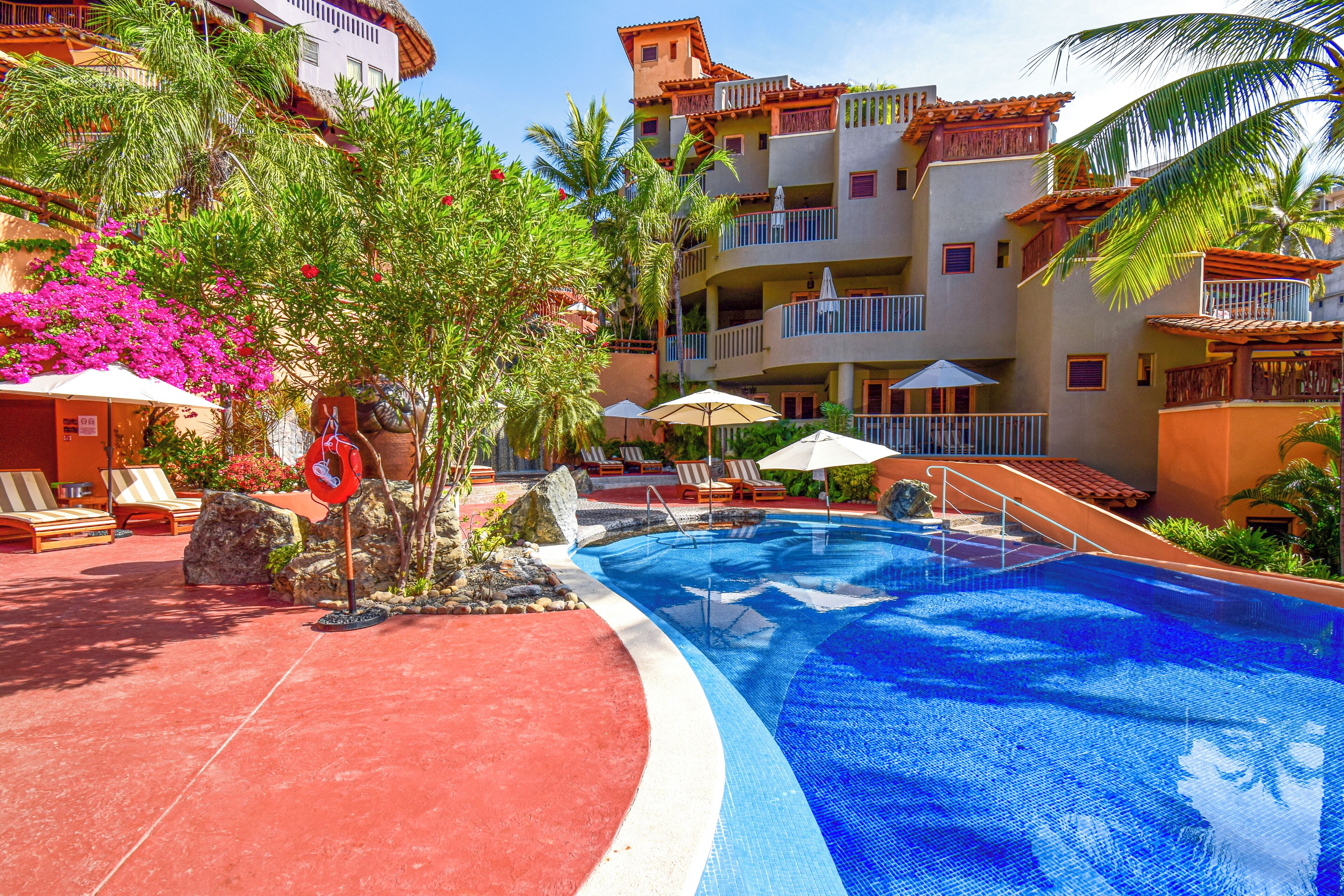 Piscina con sillones de descanso y sombrillas, rodeada de palmeras y un edificio con balcones.