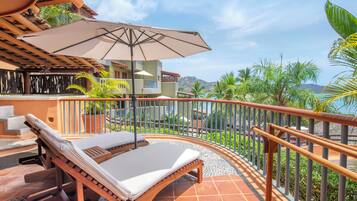 Une chaise longue avec un coussin beige et une structure en bois, placée sur une terrasse carrelée offrant une vue sur un paysage côtier et des palmiers.