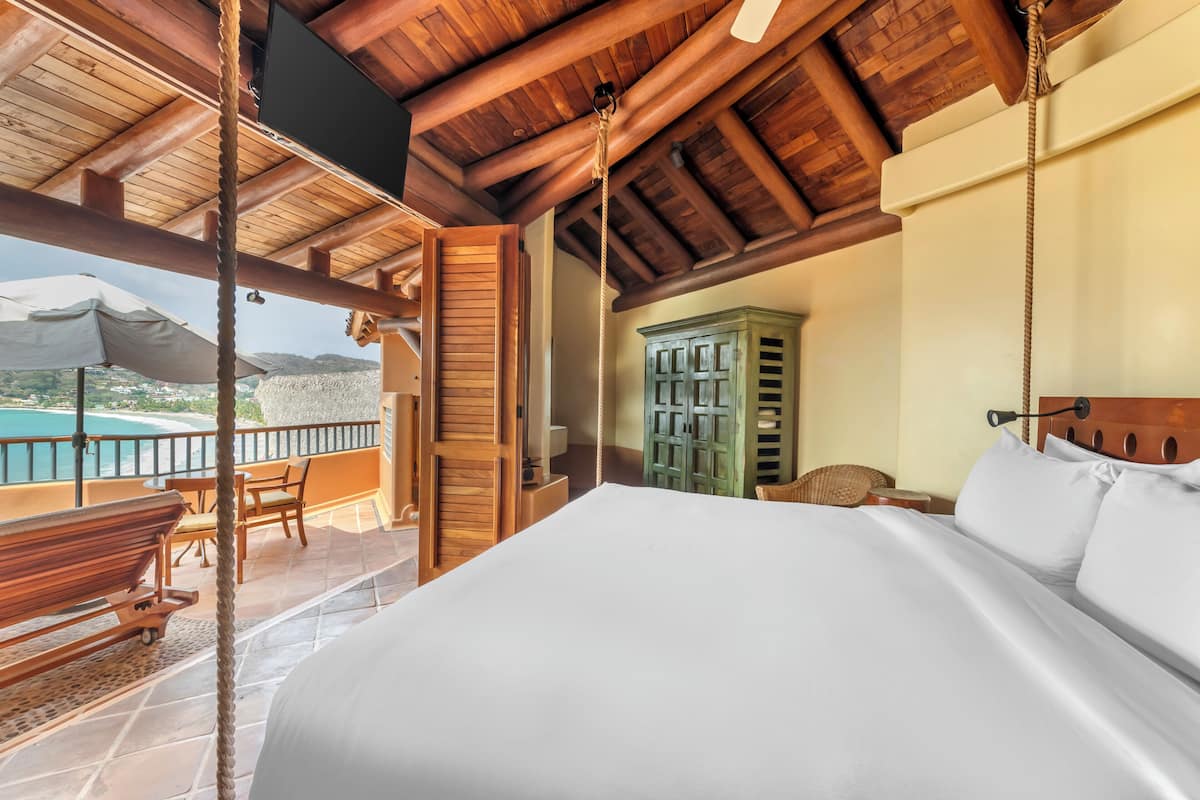 A bedroom with a large bed, wooden ceiling beams, a view of a mountain, and a balcony with a table and chairs.