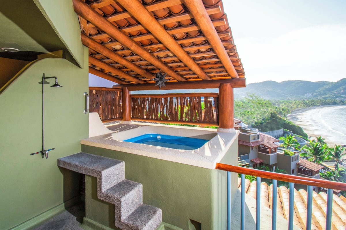 A balcony with a hot tub, a wooden pergola, and a view of a beach and mountains.