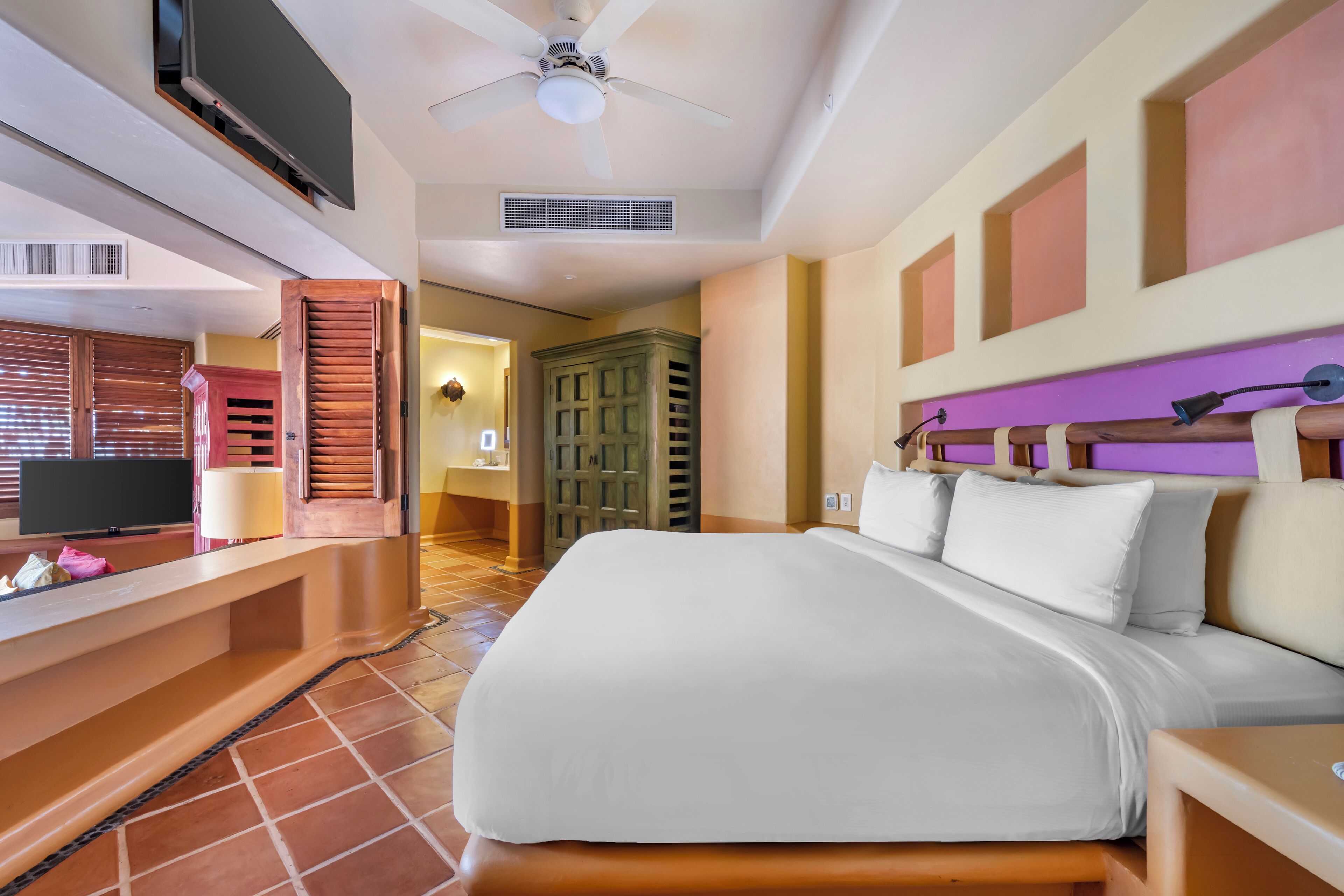 Una habitación de hotel con una cama, ventilador de techo, televisión y baño con espejo y lavabo.