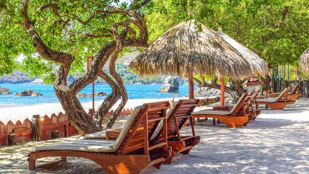 A beach with wooden lounge chairs and thatched umbrellas, a wooden fence, and a view of the sea.