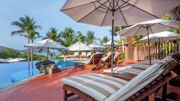 A poolside area with lounge chairs and umbrellas, surrounded by palm trees and a clear sky.
