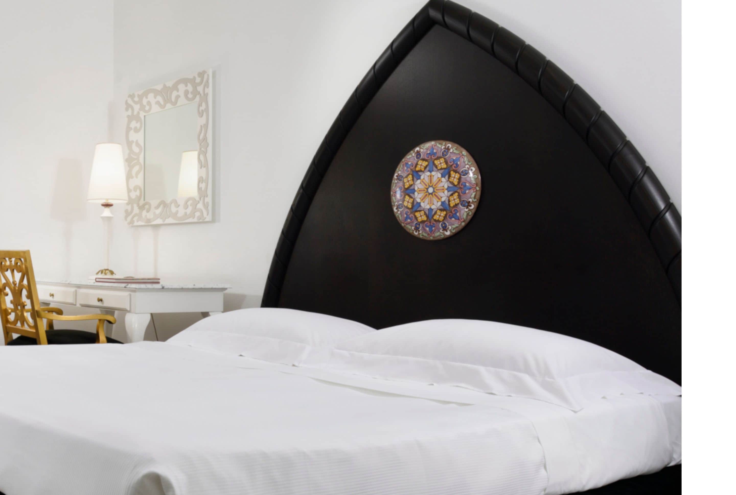 A bedroom with a black headboard, a round decorative mirror, a wooden chair, and a white bedspread.