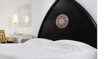 A bedroom with a black headboard, a round decorative mirror, a wooden chair, and a white bedspread.