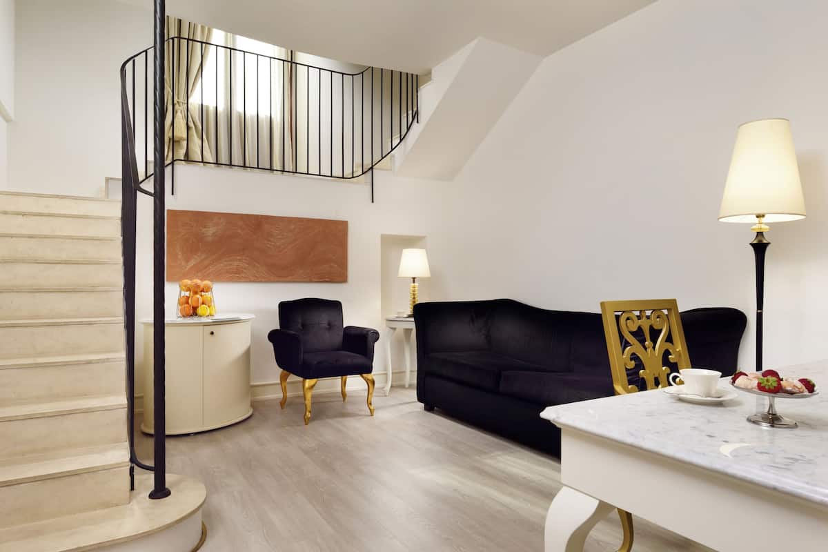 A modern living room with a staircase, a black sofa, a white table with a lamp, and a fruit bowl.