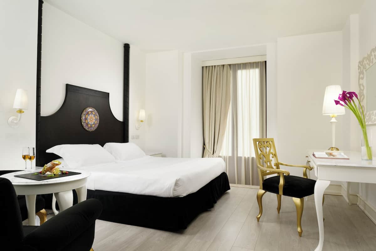 A hotel room with a bed, a desk, a chair, a small table with a lamp, and a window with curtains.