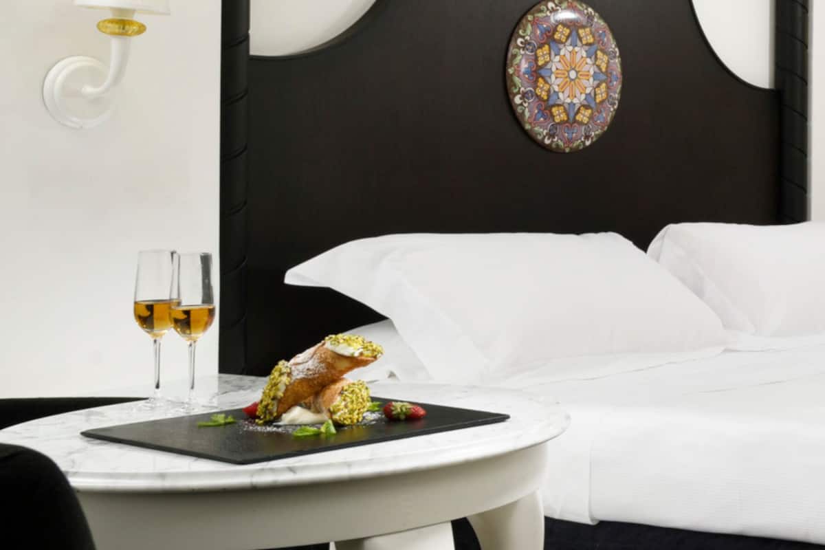 A hotel room with a bed, a bedside table with a plate of food and two glasses of wine, a decorative wall piece, and a wall-mounted lamp.