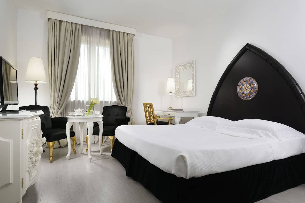 A hotel room with a large bed, a desk with a lamp, a chair, and a small table with a vase.