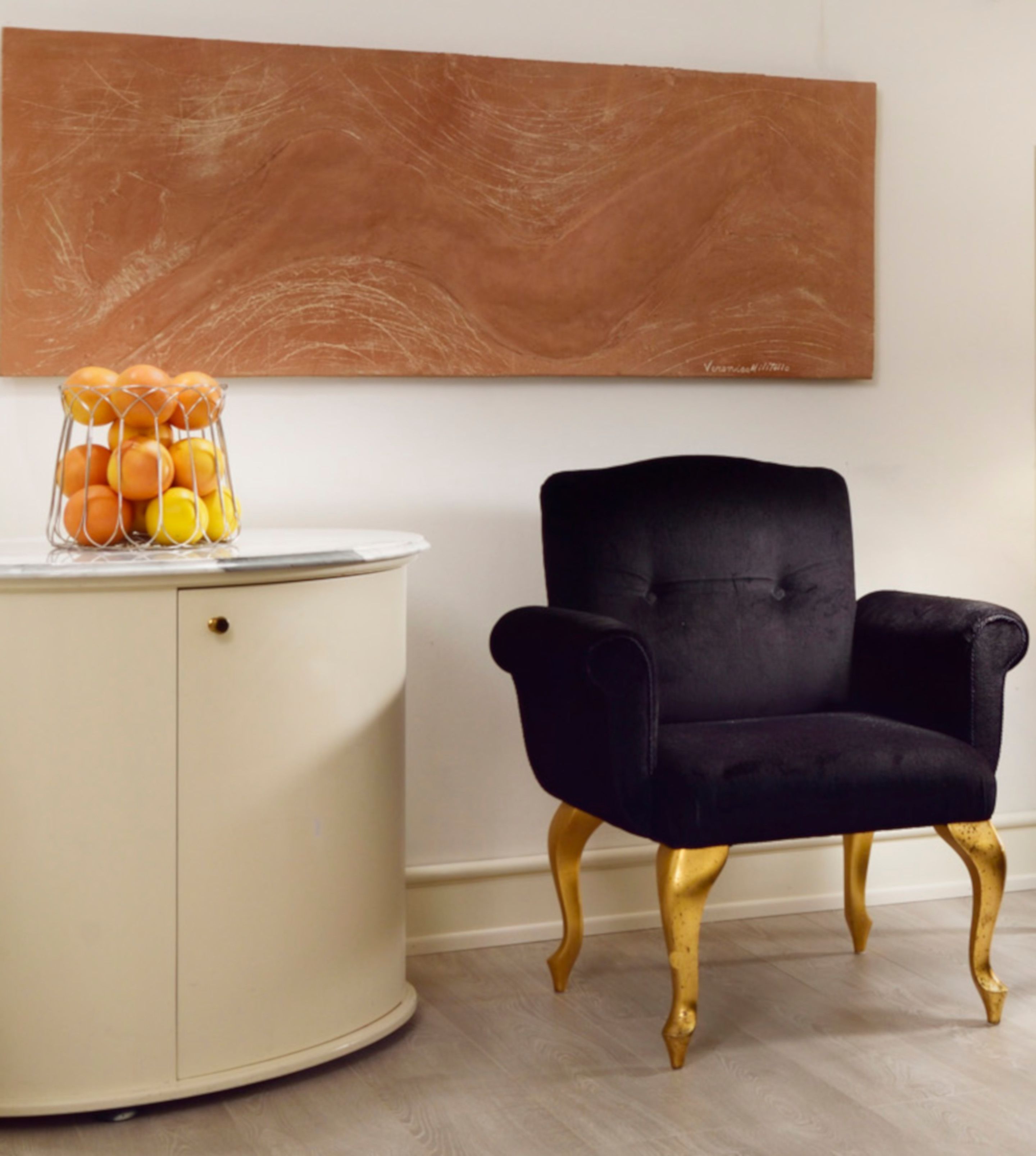 A room with a round white cabinet, a black velvet armchair with gold legs, and a large abstract painting on the wall.