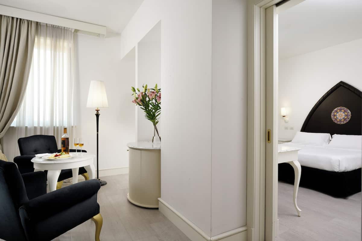 A hotel room with a bed, a desk with a lamp, a chair, a small table with a vase of flowers, and a view of another room.