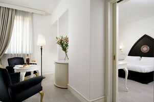 Quarto de hotel com uma cama, uma mesa com abajur, uma cadeira, uma mesinha com um vaso de flores e vista para outro quarto.