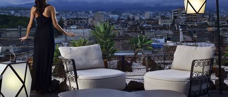 A rooftop terrace with a city view, two white cushioned chairs, and a table.